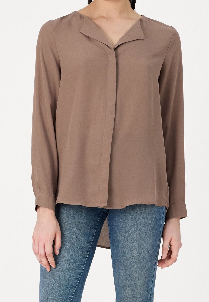 Brun langærmet bluse med v-udskæring, lavet af blødt stof. Har en knaplukning foran og en let buet kant, båret med blå jeans.