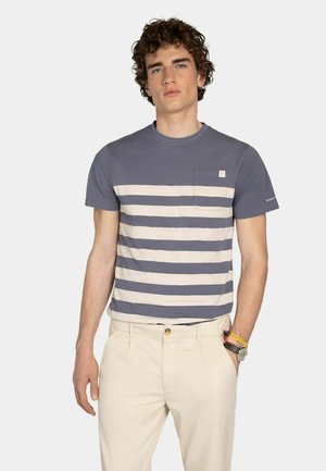 Jonge man met krullend haar, gekleed in een grijs-wit gestreept T-shirt met korte mouwen en lichtbeige broek, staand met één hand in zijn zak.