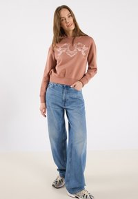 Lyse rosa sweatshirt med blomsterdesign, avslappet passform, høy krage. Kombinert med lyseblå vide jeans og mønstrede sneakers.