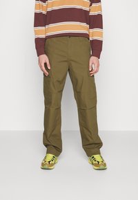 Olivgrüne Cargo-Hosen mit mehreren Taschen, geradem Schnitt, aus robustem Material, kombiniert mit gelben und braunen Turnschuhen.