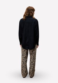 Person mit schulterlangem, welligem Haar, der ein lockeres schwarzes Langarmshirt und weit geschnittene Hosen mit Leopardenmuster trägt und mit dem Rücken zur Kamera steht.