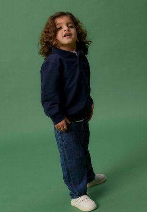 Marineblauwe hoodie met een kraag, donkerblauwe jeans en witte sneakers. Kind met krullend haar staat tegen een effen groene achtergrond.