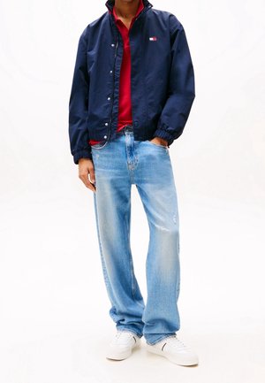 Osoba w granatowej kurtce na czerwonej koszuli, jasnoniebieskich, luźnych dżinsach i białych trampkach, stojąca z jedną ręką w kieszeni.