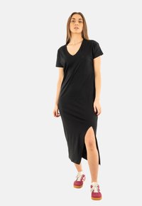 Robe noire, longueur genou, à manches courtes et décolleté en V profond. Présente une fente sur le côté et un logo discret au niveau de la poitrine. Portez-la avec des baskets colorées.