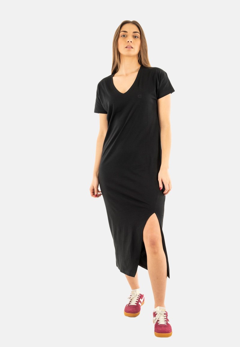 Robe noire, longueur genou, à manches courtes et décolleté en V profond. Présente une fente sur le côté et un logo discret au niveau de la poitrine. Portez-la avec des baskets colorées.