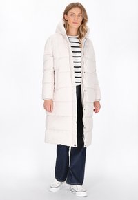 Lange, lichtroze pufferjas met horizontale stiksels, capuchon en ritsluiting. Gedeeltelijk over een gestreepte shirt gedragen en gecombineerd met donkere blauwe jeans.