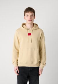 Sudadera capucha beige con un bolsillo frontal y un parche de logo rojo en el cuello. Tejido suave, mangas largas y puños acanalados.