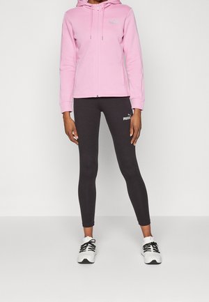 Personne portant un sweat à capuche rose zippé, des leggings Puma noirs et des chaussures de sport blanches, debout devant un fond uni.