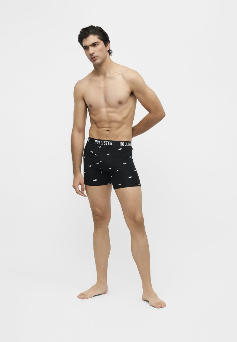Гол мъж, стоящ бос, облечен в черни боксерки Hollister с лого на колана и малки бели птици на бял фон.