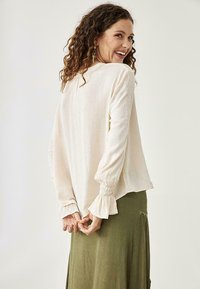 Blusa de mangas largas en beige claro con corte holgado, puños fruncidos y textura suave, combinada con una falda verde oliva con acentos decorativos.