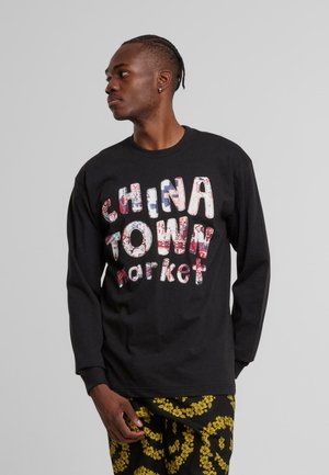 Svart langermet t-skjorte med "CHINA TOWN market" i fargerike, teksturerte bokstaver. Sammenkoblet med mønstrede svarte bukser.