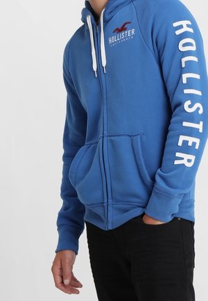 Blauer Zip-Hoodie mit weißem Text „"Hollister"" auf Ärmel und Brust, getragen von einer Person mit einer Hand in der Tasche der schwarzen Hose.