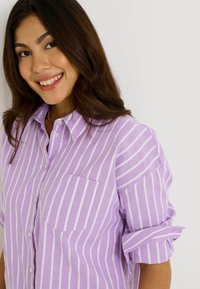 Camisa de botones a rayas lavanda con un bolsillo en el pecho, que presenta rayas verticales blancas y mangas enrolladas. Material de algodón o lino.