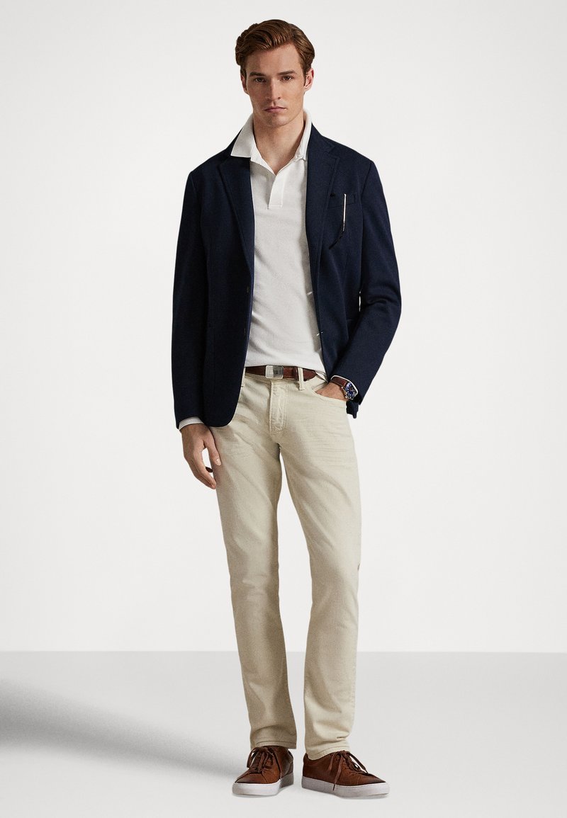 Homme portant un blazer bleu marine sur un polo blanc, pantalon beige, ceinture marron, baskets marron, debout avec une main dans la poche sur un fond uni.