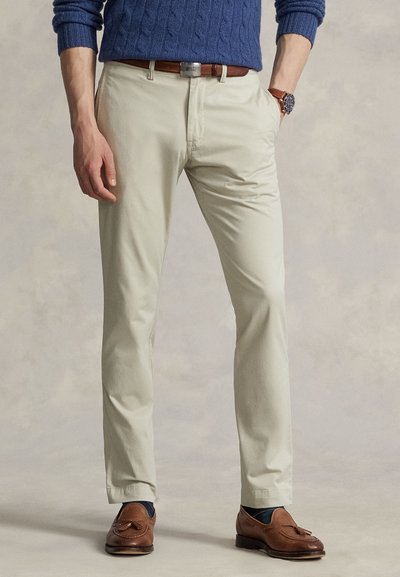 Moški, oblečen v bež chino hlače z rjavim pasom, modri pulover, rjavi loaferji s cofki in ročno uro, stoji z rokami delno v žepih.