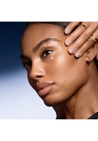 Närbild av ett ansikte med slät, strålande hud, mörkt hår, tre vita prickar nära ögat och subtilt glansiga läppar. Guldringar pryder fingrarna.