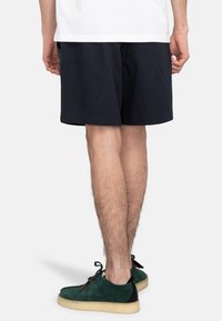 Svarta bomullsshorts med en avslappnad passform, med en enkel design. Personen har på sig gröna mockaskor med kontrasterande svarta detaljer.