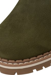 Olajzöld bőrbakancs kerek orral, kontrasztos barna gumitalppal, varrott díszítéssel és texturált felületi részletekkel.