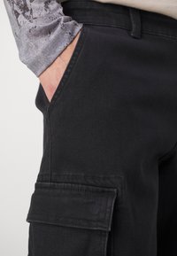Pantalones cargo negros hechos de una tela duradera, con bolsillos laterales y un diseño ajustado, con una camisa de manga larga gris texturizada parcialmente visible.