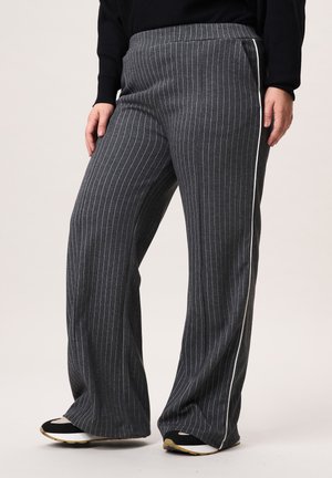 Persoon die donkergrijze wijde gestreepte broek draagt met witte strepen aan de zijkanten en zwart-witte sneakers, staand op een witte vloer.