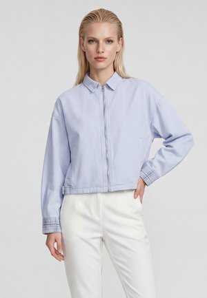 Blonde Frau mit glatten Haaren, die eine hellblaue Reißverschlussjacke und weiße Hose trägt, steht mit einer Hand auf der Hüfte vor einem einfarbigen Hintergrund.