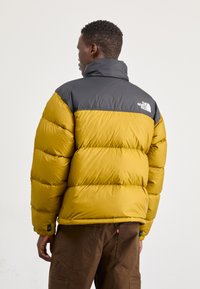 Geel pufferjack met grijze schouders, voorzien van een hoge kraag, gestikte panelen en zwarte branding op de achterkant. Glad oppervlak.