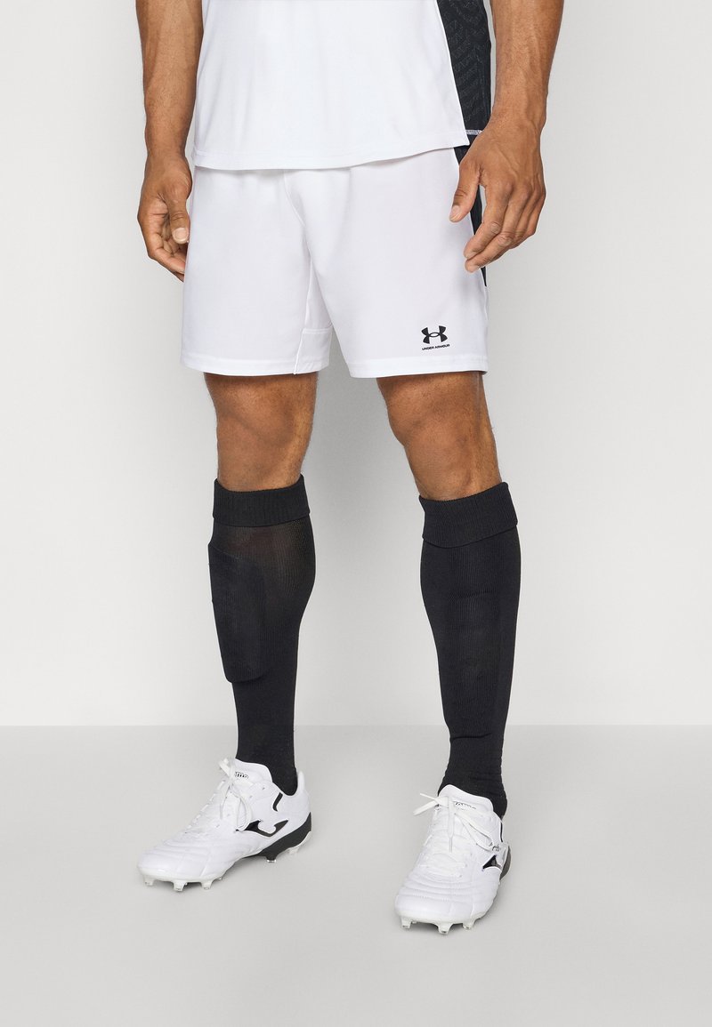 Weiße Sportshorts mit schwarzem Under Armour-Logo, kombiniert mit knielangen schwarzen Socken und weißen Fußballschuhen mit schwarzen Akzenten.