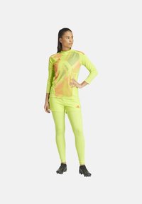 Neon gele sportoutfit met een lange mouwen top met geometrische patronen en bijpassende strakke broek. Zwarte voetbalschoenen maken de look af.