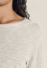 Svetlosivý, štruktúrovaný pletený pulóver so širokým výstrihom a viditeľným šitím. Mäkká látka jemne splýva cez plece.