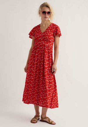 Vrouw in een rode midi-jurk met bloemenprint, bruine sandalen en rode zonnebril, staand tegen een lichte neutrale achtergrond.