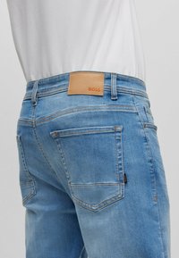 Světle modré džínové kalhoty s hnědou koženou nášivkou s logem „BOSS“ na zadní straně, mají pět kapes a kontrastní švy.