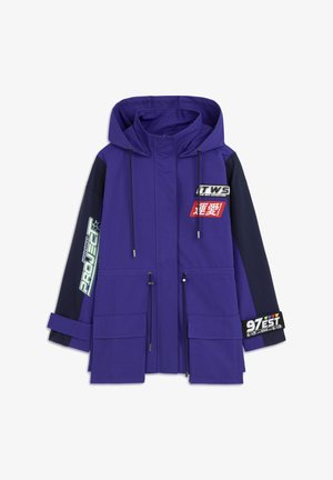 Veste d'extérieur violette et marine avec capuche, cordons de serrage, plusieurs poches et divers patchs colorés portant des textes et des graphiques.