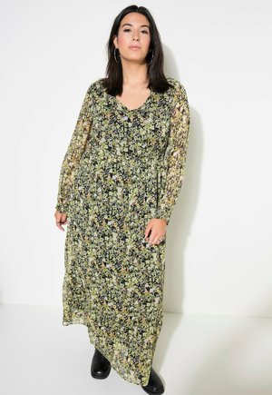Zwarte maxi-jurk met bloemenprint in groen en oranje, lange doorschijnende mouwen en een V-hals. Gecombineerd met zwart schoeisel.