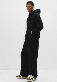 Junger Mann trägt einen schwarzen übergroßen Kapuzenpullover und weite schwarze Hosen, steht mit einer Hand in der Tasche und grauen Sneakers mit weißen Schnürsenkeln.