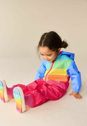 Fiatal, sötét hajú gyermek színes esőkabátban és rózsaszín, szivárványos esőcsizmában ül a padlón, lefelé néz és mosolyog.