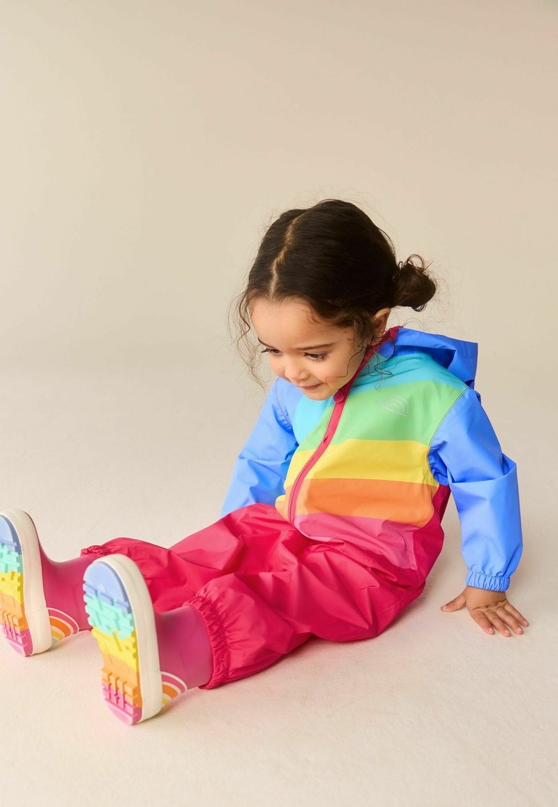Copil tânăr cu păr închis la culoare, purtând o pelerină de ploaie colorată și cizme de ploaie roz cu curcubeu, stând pe podea, uitându-se în jos și zâmbind.
