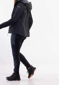 Sort udendørsjakke med hætte, lynlåslommer; parret med sorte bukser med teksturerede paneler og sorte sneakers med orange detaljer.