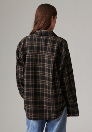 Personne aux longs cheveux bruns portant une chemise à carreaux noire et marron à manches longues et un jean bleu, de dos sur un fond uni.