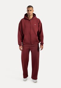 Sudadera de color burdeos con cremallera, con un bolsillo frontal y pantalones sueltos a juego. Hecha de una tela suave, cuenta con un pequeño logo en el pecho.
