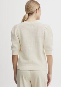 Kremfarget strikket genser med teksturert stoff og puffede ermer. Ribbekanter på ermer og nederkant fremhever designet. Sett fra baksiden.