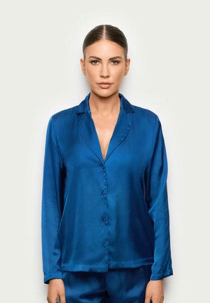 Mujer con el cabello recogido que lleva una camisa azul brillante abotonada de manga larga y pantalones a juego, de pie contra un fondo liso y claro.