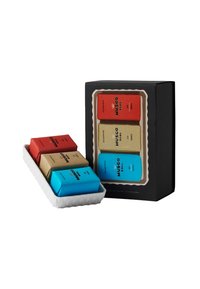 Geschenkset mit drei Seifen, verpackt in rotem, beigem und blauen Papier, präsentiert in einer weißen, gewellten Schale, mit einer schwarzen Verpackungsbox.