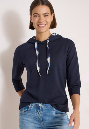 Smilende kvinde iført en mørkeblå hættetrøje med mønstrede snore og lyseblå jeans mod en ensfarvet lys baggrund.