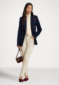 Tengerészkék dupla soros blézer arany gombokkal, krémszínű ing megkötős nyakkal, bézs nadrág, bordó kézitáska és hozzá illő bordó mokaszinok.