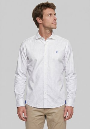 Hombre de pie frente a un fondo gris, vistiendo una camisa blanca abotonada con logotipo azul y pantalones beige, mirando hacia arriba a su derecha.