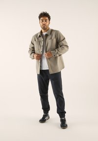 Veste en flanelle beige en laine avec deux poches poitrine, boutons noirs et coupe décontractée, portée sur un t-shirt blanc et un pantalon bleu marine.