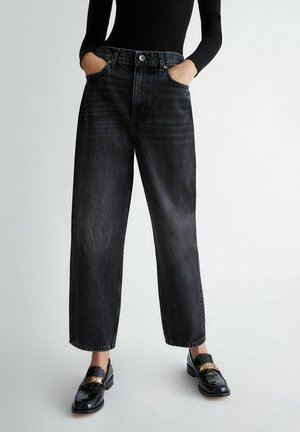 Henkilö pukeutuneena korkeavyötäröisiin haalistuneisiin mustiin leveälahkeisiin farkkuihin, kädet taskuissa, ja kiiltäviin mustiin loafer-kenkiin, joissa on kullanvärisiä yksityiskohtia.