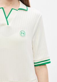 Λευκό ριμπ πικέ πόλο με πράσινη επένδυση και γιακά, με στρογγυλό λογότυπο σε πράσινο και λευκό στο στήθος. Κοντά μανίκια με ρίγες.