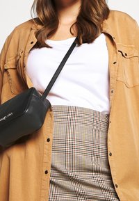 Czarna torebka crossbody, beżowa koszula, a na to biała koszulka na kratkowej spódnicy z żółtymi i brązowymi wzorami. Gładka tekstura i dopasowany fason.