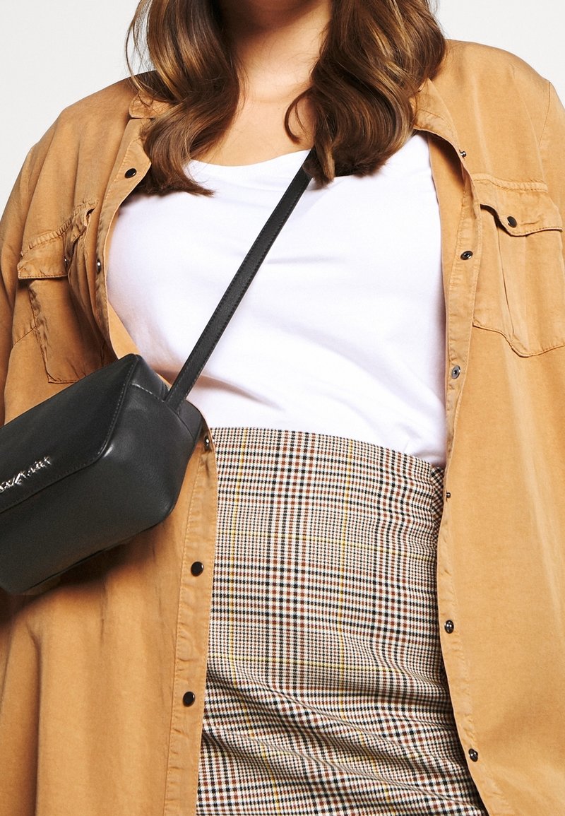 Czarna torebka crossbody, beżowa koszula, a na to biała koszulka na kratkowej spódnicy z żółtymi i brązowymi wzorami. Gładka tekstura i dopasowany fason.